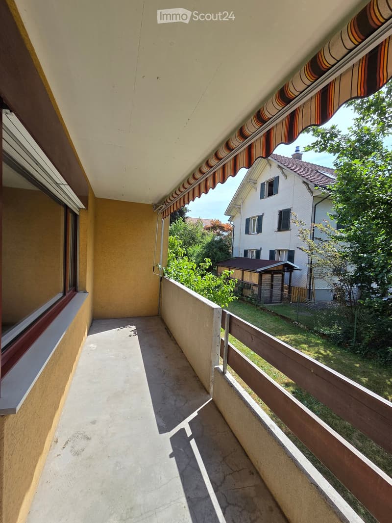 Balcony with a striped awning, wooden railing, and view of a garden and neighboring house.