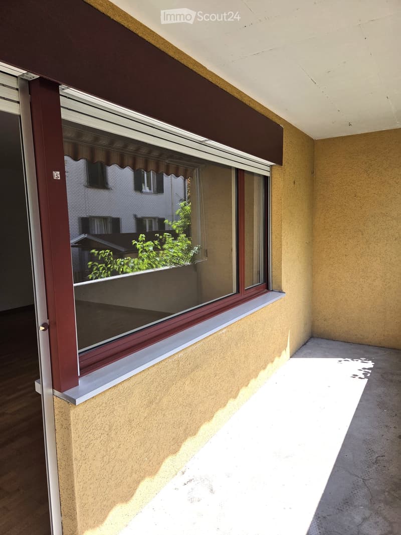 Balcony with large window, beige wall, and view of neighboring building and greenery.