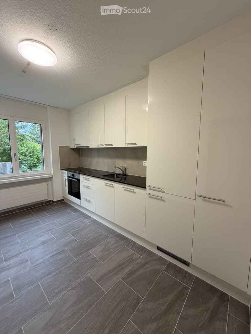 Modern kitchen with white cabinets, dark countertops, and a stainless steel sink. Features include a built-in oven and a large window with a view of greenery.