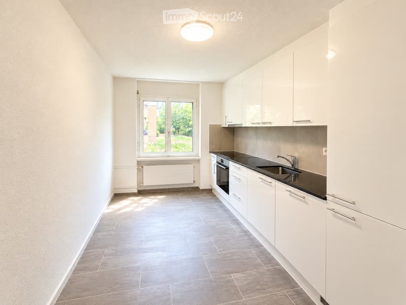 Modern kitchen with white cabinets, dark countertops, and a built-in oven. Features a large window with a view of greenery outside.