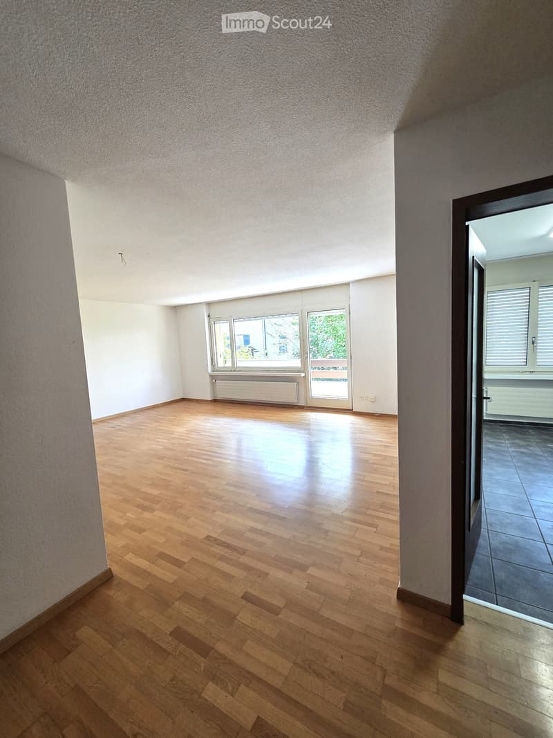 Empty room with wooden flooring, white walls, large windows, and a door leading to another room.