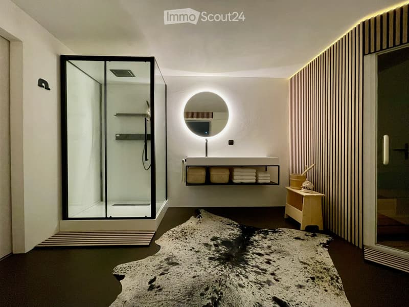 Modern bathroom with a large, illuminated round mirror above a sleek sink, a glass-enclosed shower, and a small wooden stool with towels and toiletries.