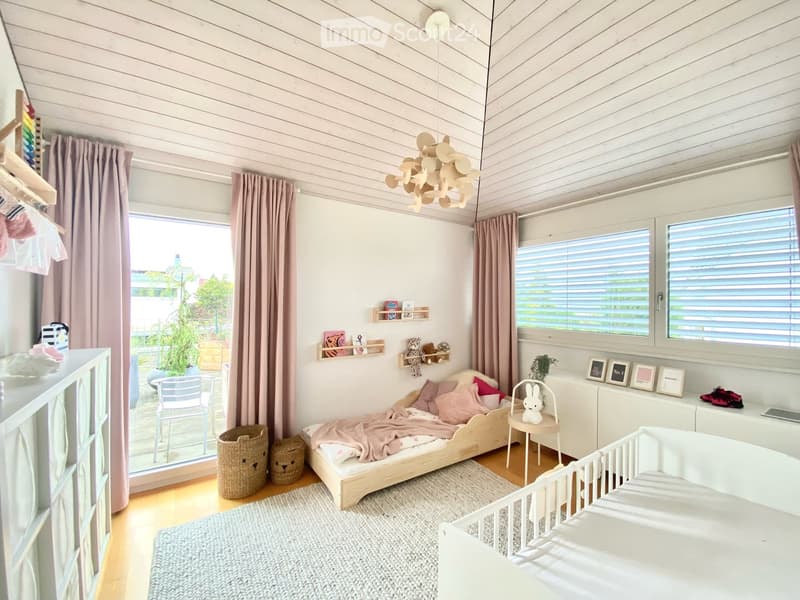 Bright, modern children's bedroom with a crib, daybed, small table, and shelves. Features large windows with blinds, a hanging wooden lamp, and a cozy rug.