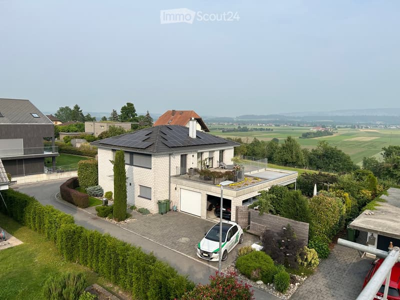 A modern two-story house with solar panels on the roof, a garage, and a well-maintained garden. The driveway has a parked car, and the surrounding area features open fields and trees.