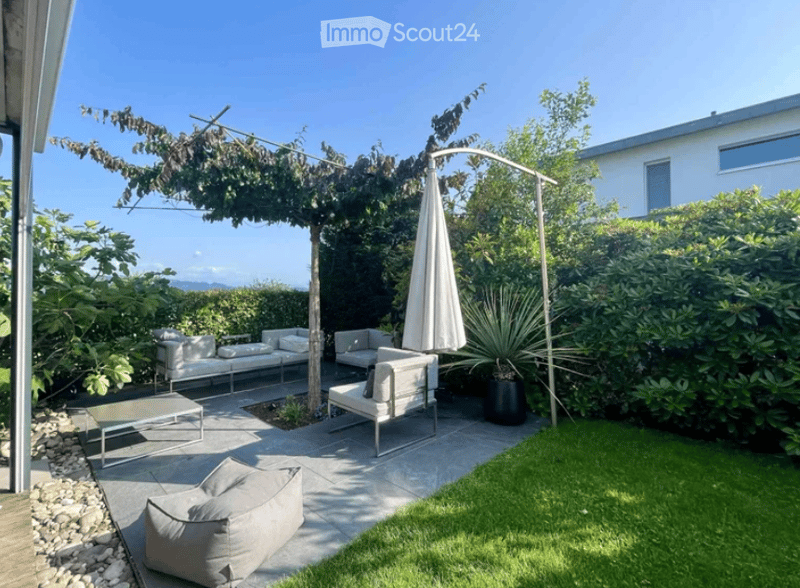 Modern outdoor patio with white furniture, including a sofa, chairs, and a coffee table, surrounded by greenery and a pergola with an umbrella.