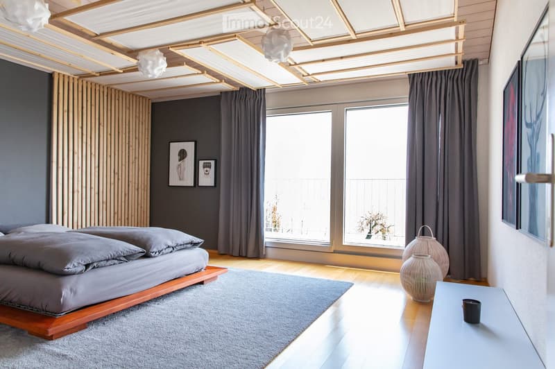 Modern bedroom with a wooden bed, nightstand, wicker lamps, and large windows with curtains. The room features a grey duvet, grey pillows, and a grey rug on the floor.