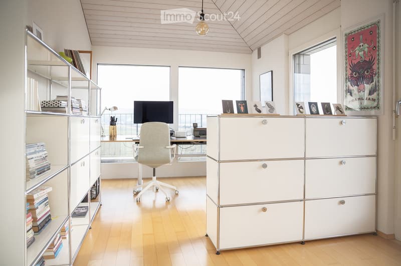 Bright, modern office space with wooden flooring, white walls, and large windows. Features a white desk, ergonomic chair, shelving unit with books, and a modular storage unit.
