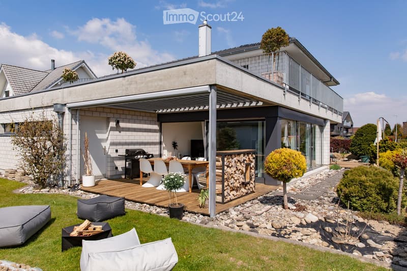 Modern outdoor patio with wooden deck, dining table and chairs, grill, fire pit, bean bags, and potted plants.