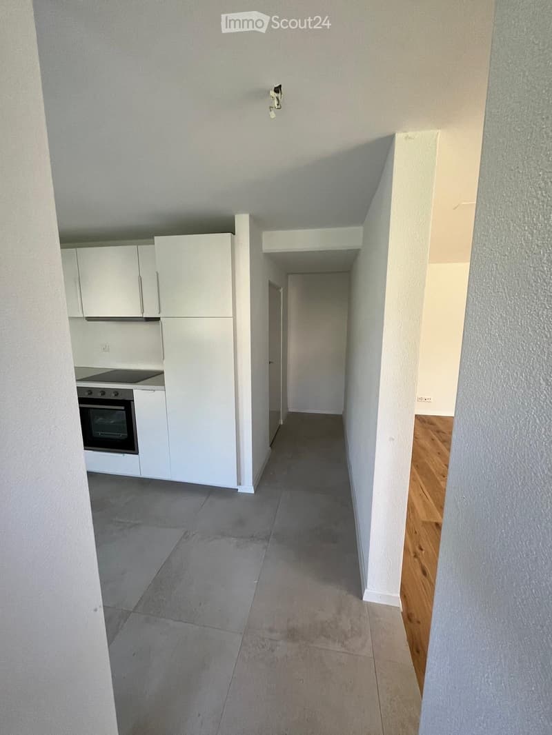 Empty hallway with grey tiled floor, white walls, and wooden flooring visible in the adjacent room. Kitchen area visible on the left with white cabinets, stove, and refrigerator.