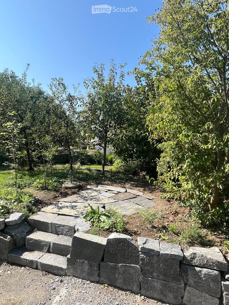 Garden area with a stone pathway, stone wall, and surrounding trees and shrubs.