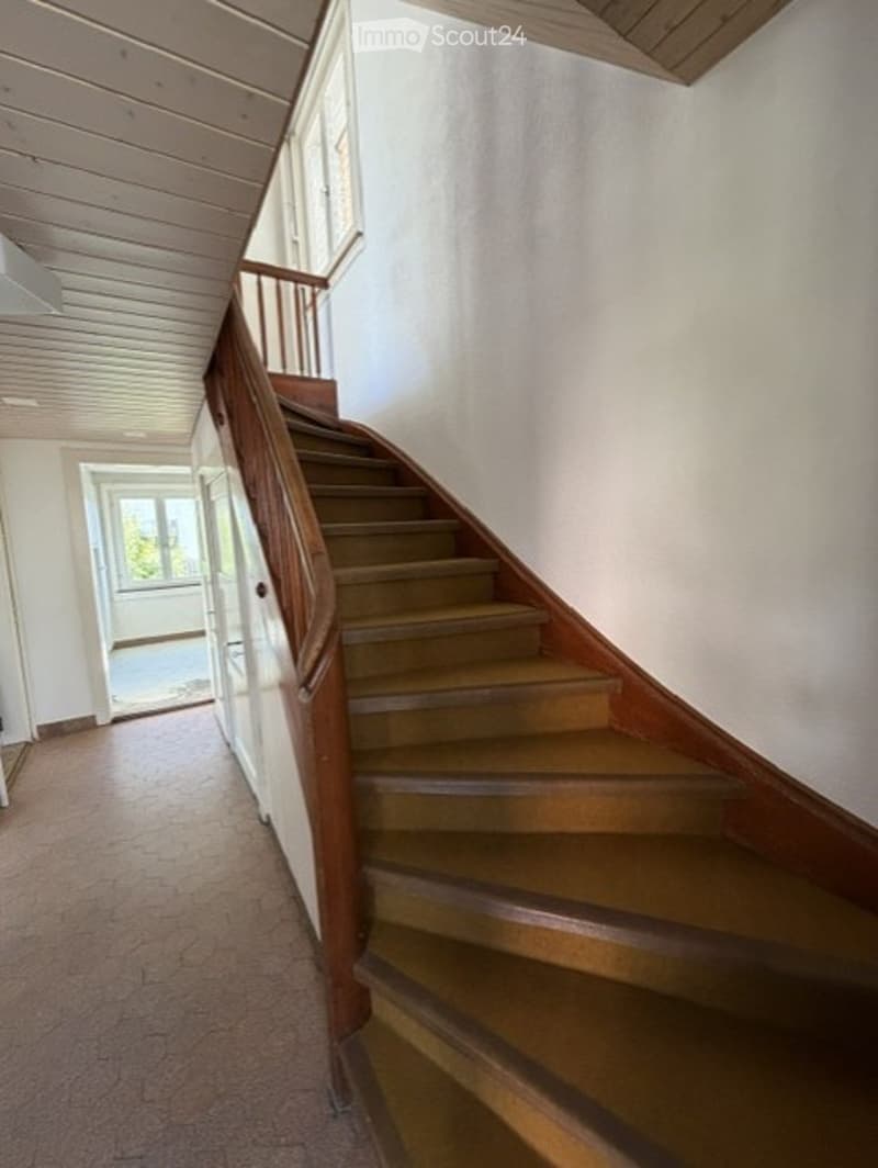 Spacious hallway with wooden staircase, light-colored walls, and carpeted floor. The staircase features wooden railings and steps, leading upwards. Natural light filters in from a window at the top of the stairs.
