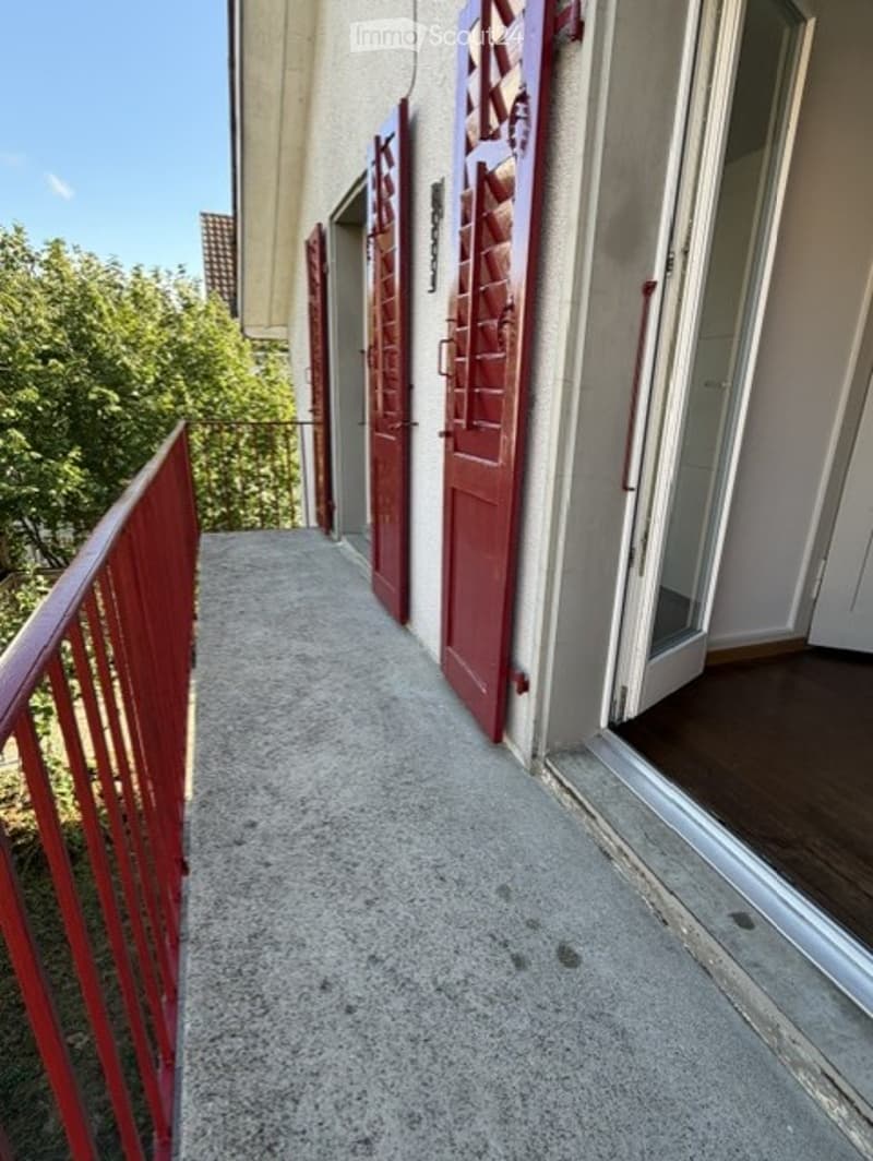 A narrow concrete hallway with red metal railings on both sides, leading to several closed doors with decorative red shutters.