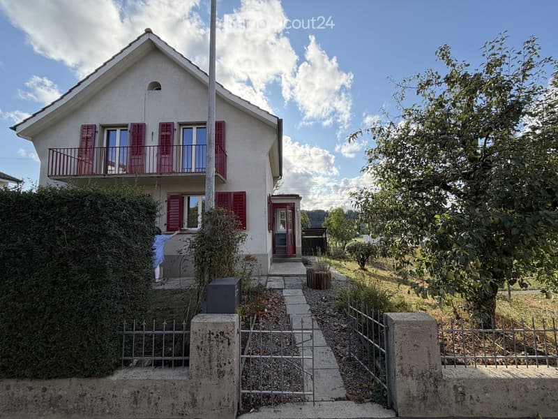Two-story house with white exterior, red shutters, and a balcony on the upper floor. The house is surrounded by a garden with trees and a pathway leading to the entrance.