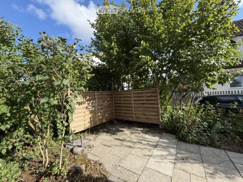 Private garden area with wooden fence, paved patio, trees, and shrubs.