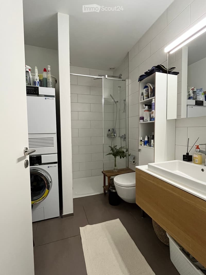 Modern bathroom with a white tiled shower, wooden vanity, sink, toilet, washing machine, and shelves holding cleaning supplies.