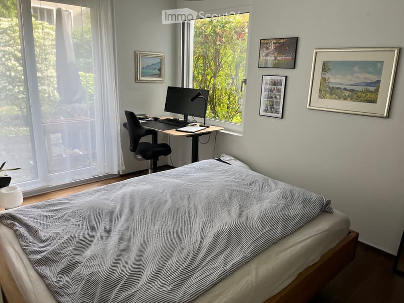 Cozy bedroom with a double bed, desk with a laptop and chair, large windows with sheer curtains, and several framed pictures on the wall.