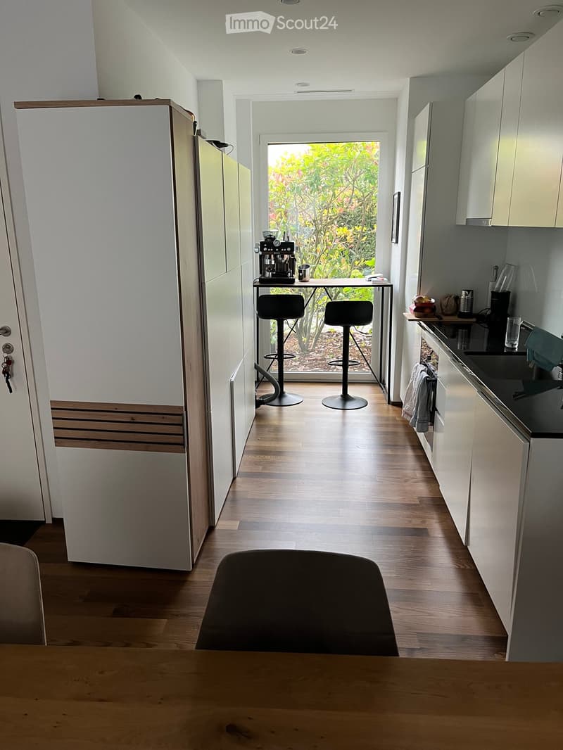 Modern kitchen with light-colored cabinets, wooden flooring, and a bar area with stools. Features a coffee machine, sink, and refrigerator.