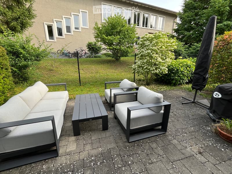 Outdoor patio with grey sofa, two chairs, and a coffee table, surrounded by greenery and a covered umbrella.