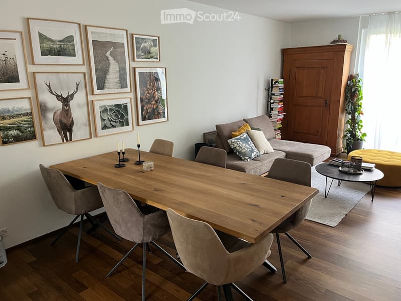 A modern living and dining area with a wooden dining table, six chairs, a grey sofa with pillows, an ottoman, a coffee table, and several framed pictures on the wall.