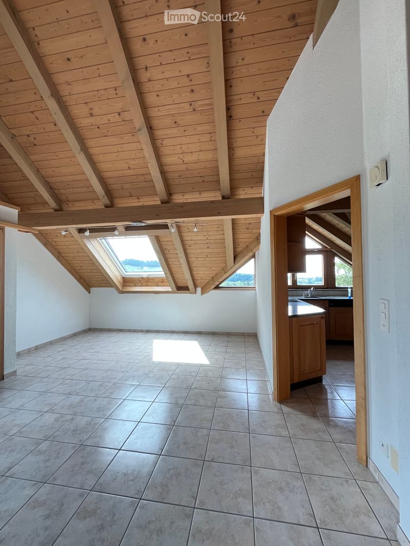 Empty room with exposed wooden beams and skylight, tiled floor, and a small kitchen area visible through an open doorway.