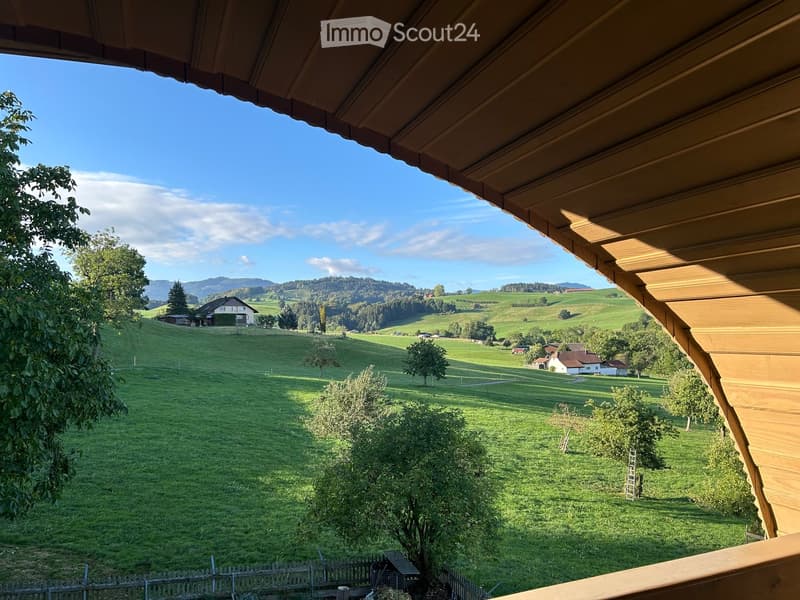 A scenic view of a lush green valley with rolling hills, scattered trees, and a few houses in the distance under a clear blue sky.