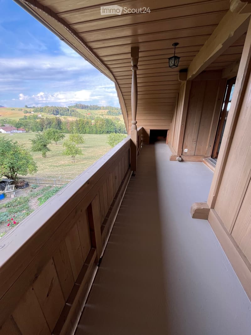 Wooden balcony with a railing, wooden ceiling, and a view of a rural landscape with fields and trees.