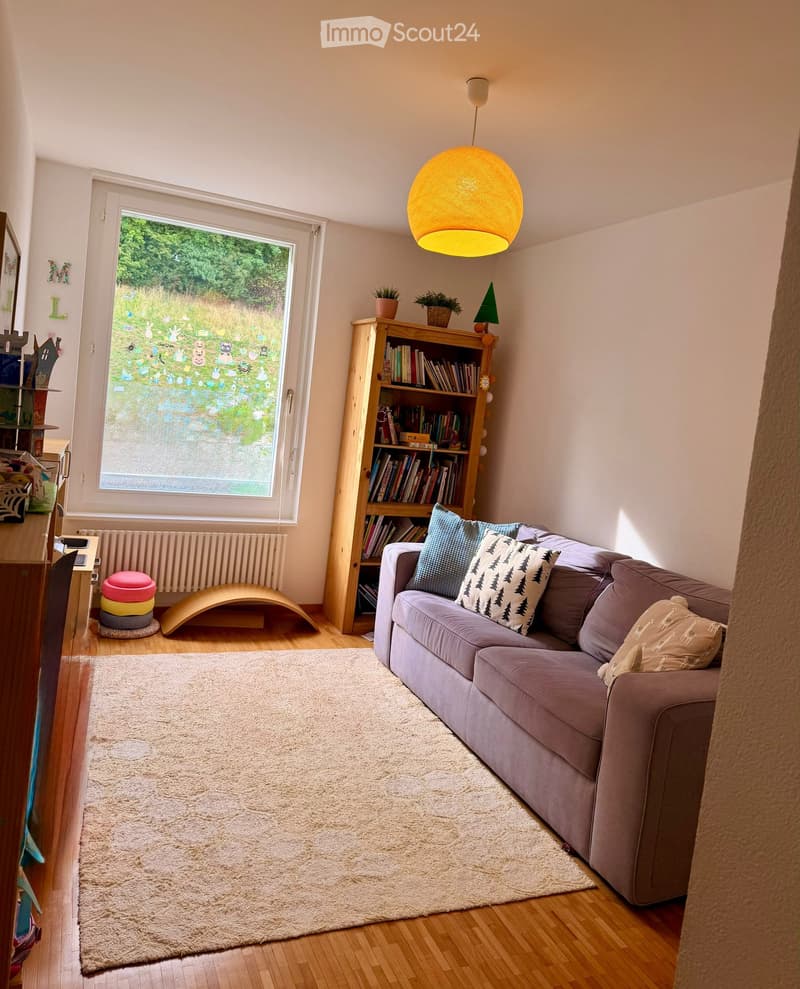 Living room with a grey sofa, bookshelf, potted plants, floor lamp, and a beige rug on wooden flooring.