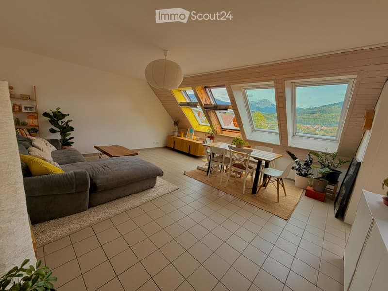 Bright living room with large windows offering a scenic mountain view, featuring a grey sectional sofa, coffee table, dining area with a table and chairs, and several potted plants.