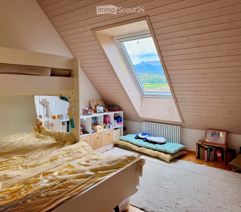 Cozy attic bedroom with wooden slanted ceilings, skylight window offering a scenic mountain view, a large bed with yellow bedding, a small seating area with cushions, built-in storage, and a bookshelf.