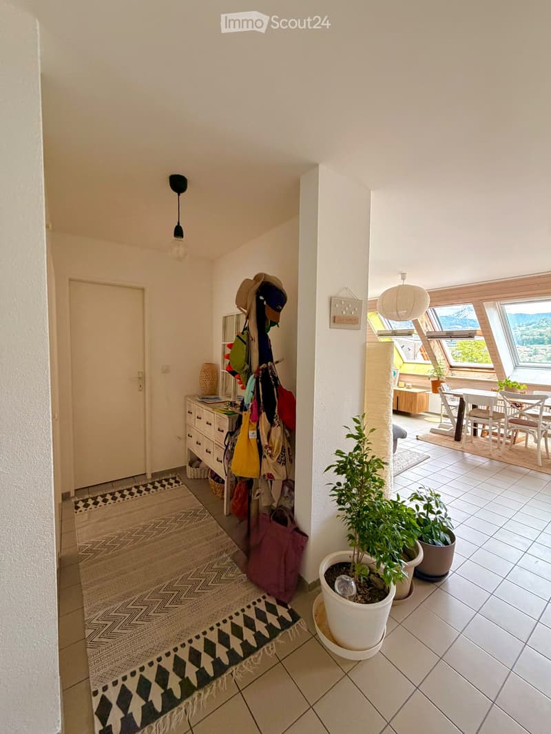 Bright hallway with tiled floor, white walls, and a view to a dining area with a table and chairs. Features a coat rack with bags and hats, a small cabinet, and potted plants.
