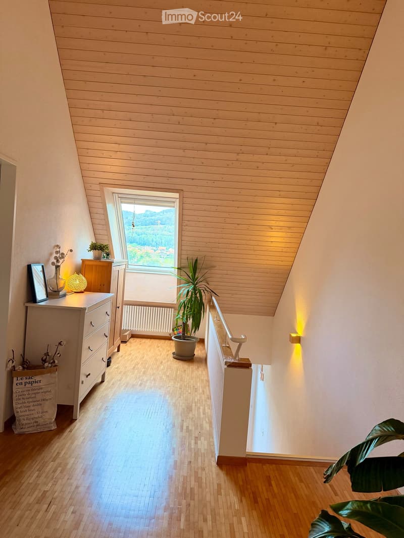 Bright hallway with wooden floors and paneled ceiling, featuring a large window with scenic view, small table with lamp and plant, and a potted plant on the floor.