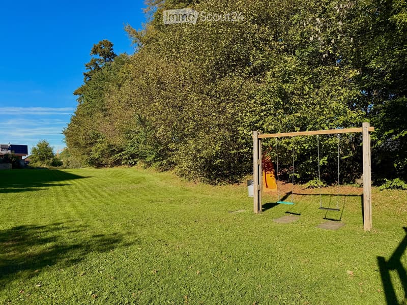 A well-maintained garden with a swing set featuring swings and a slide, surrounded by lush green trees and grass under a clear blue sky.