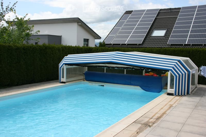 A swimming pool with a covered patio area, surrounded by a hedge and a modern house with solar panels on the roof.