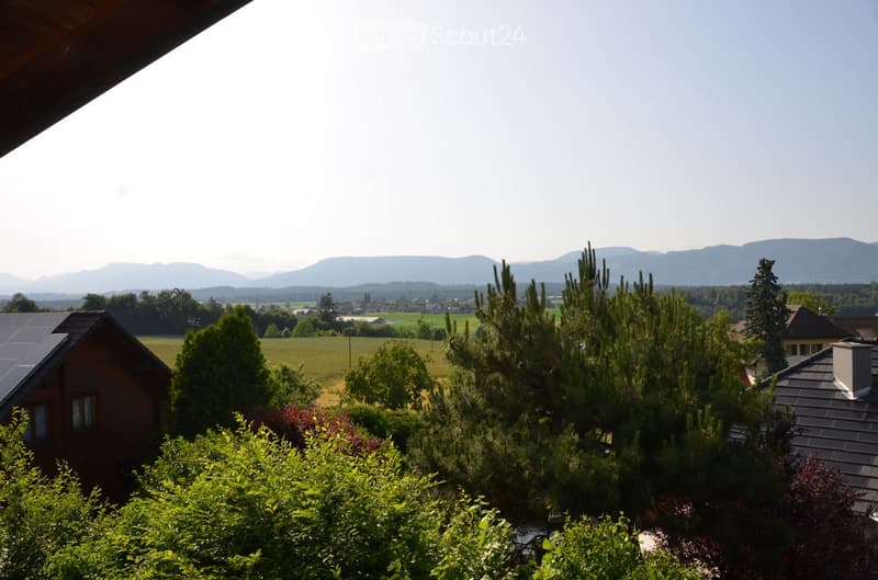 A serene garden view with lush greenery, including bushes and trees, overlooking a picturesque countryside landscape with fields, scattered houses, and distant mountains under a clear sky.