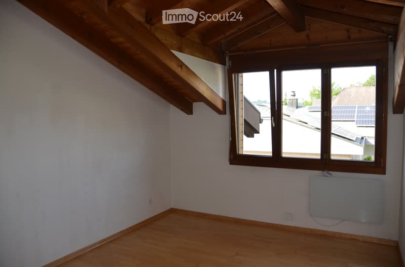 Empty attic room with wooden beams, wooden floor, white walls, and a large window overlooking a rooftop with solar panels.