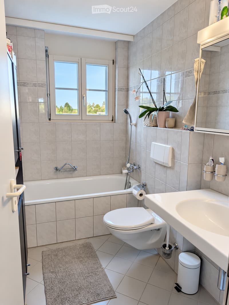 Modern bathroom with a bathtub, toilet, sink, and mirror. Features tiled walls and floor, a window with blinds, and a small plant on the shelf.
