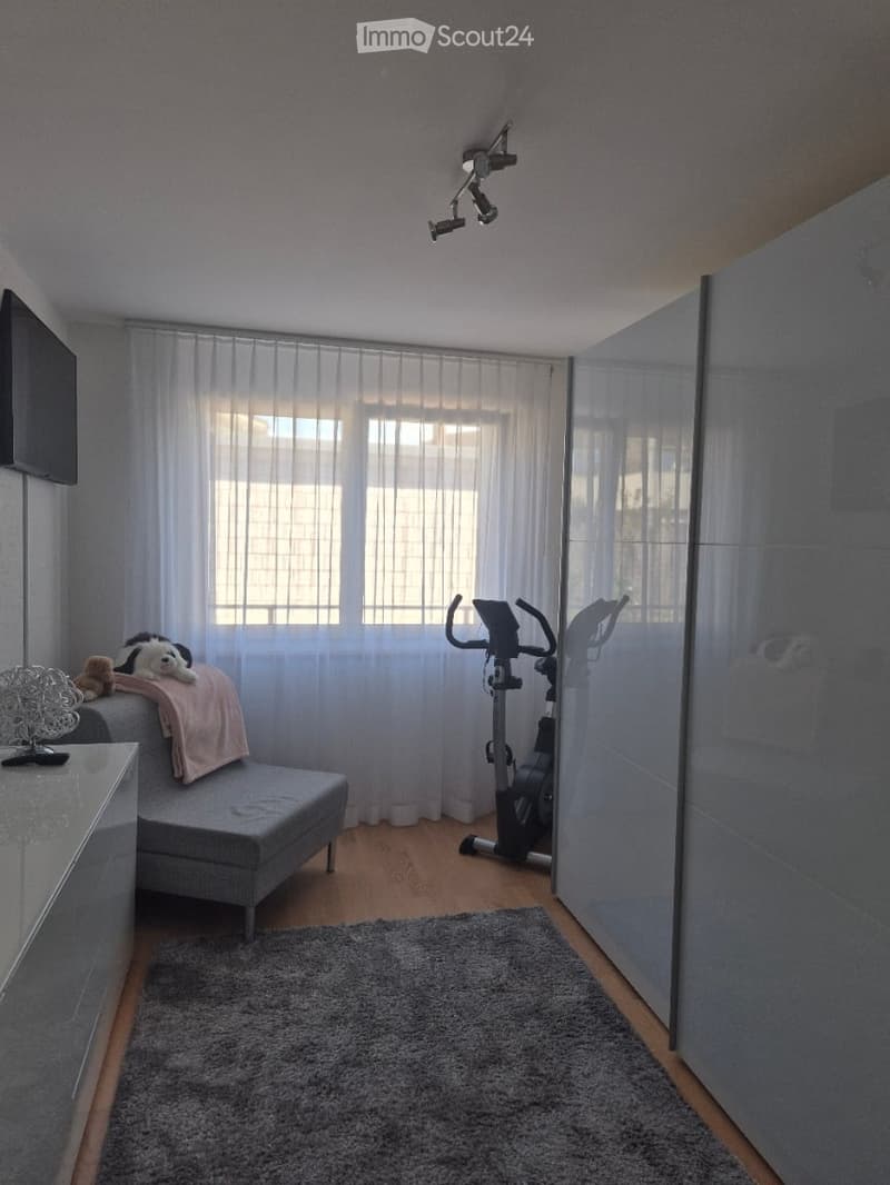 A modern living room with a grey sofa, exercise bike, large window with white curtains, and a small table with a lamp and decorative items.