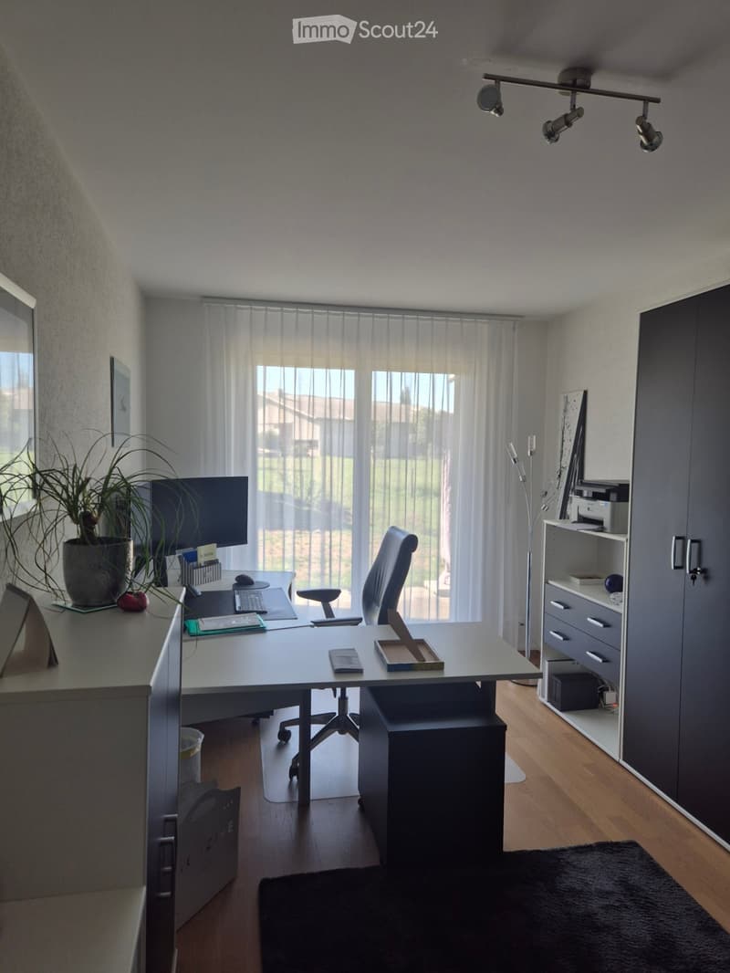 Modern office space with a large desk, ergonomic chair, computer setup, and shelving unit. Features large windows with sheer curtains, wooden flooring, and contemporary lighting.