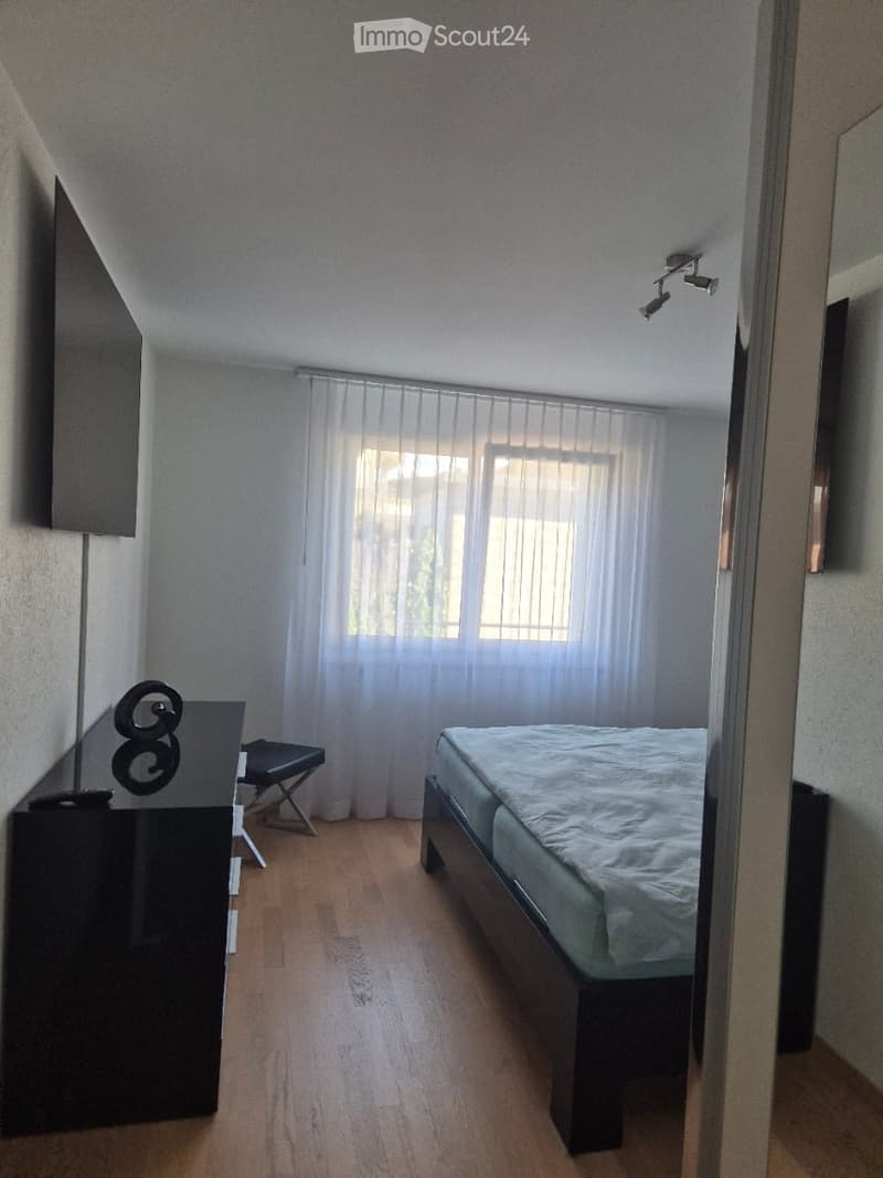 Modern bedroom with wooden flooring, a double bed with white bedding, a black nightstand with a lamp, a small black stool, and sheer white curtains covering a window.