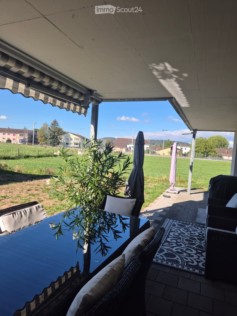 A covered outdoor patio with a glass table, wicker chairs, and an umbrella, overlooking a grassy field and houses.