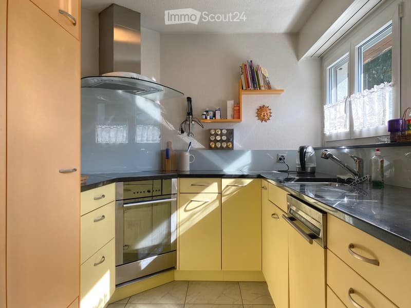 Modern kitchen with light-colored cabinets, stainless steel appliances, and a small window with a curtain. Includes a faucet, oven, microwave, refrigerator, and a small bookshelf with books.