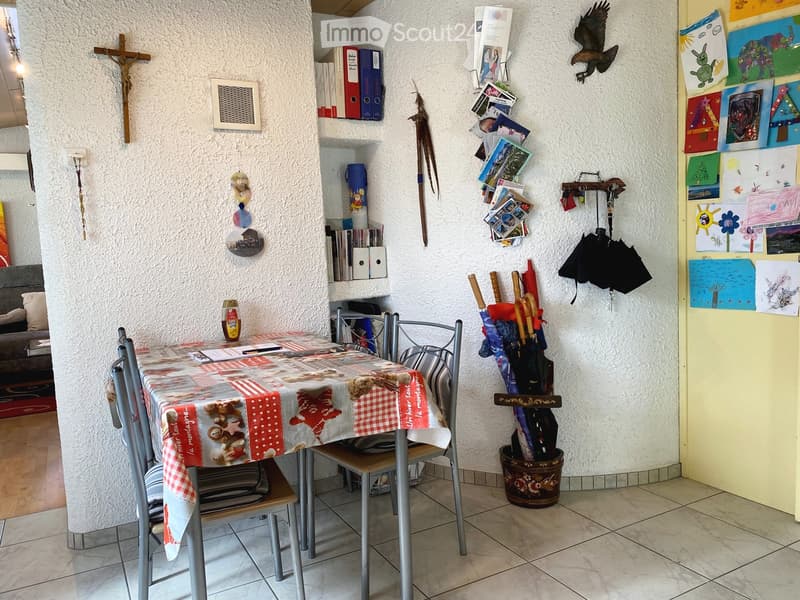 A narrow hallway with white walls, decorated with various items including umbrellas, artwork, books, and a small shelf with storage compartments. The hallway features a staircase leading upwards and a small table with chairs in the background.