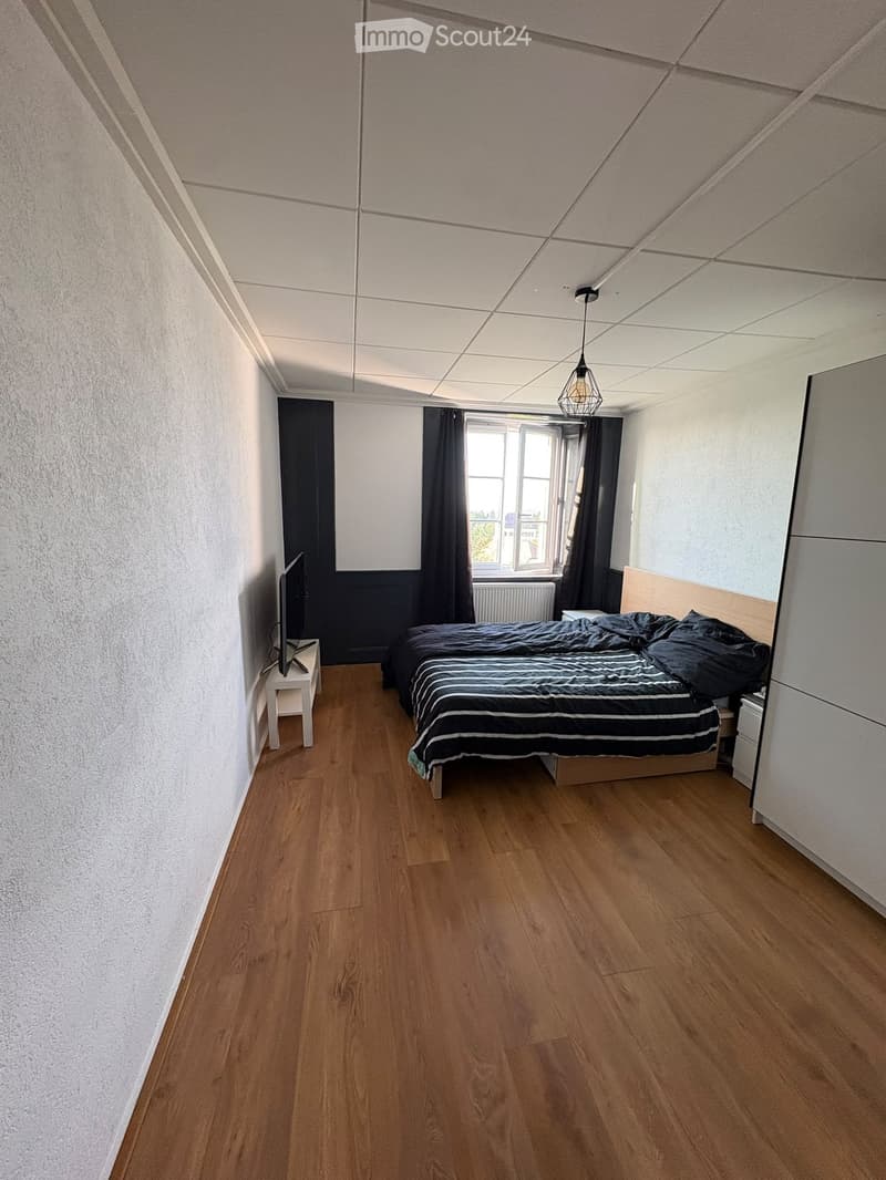 Bedroom with wooden flooring, a double bed with black and white striped bedding, a nightstand, a television on a small table, and dark curtains on the windows.