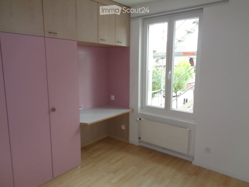 Empty room with light pink walls, wooden floor, built-in cabinets, and a window with a view of the street.