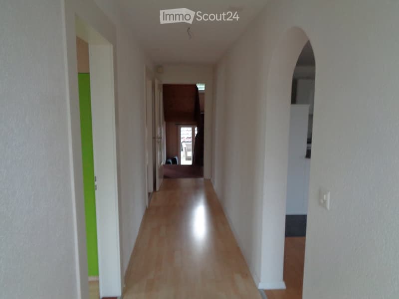 A long, narrow hallway with light wooden flooring and white walls. The hallway features several doors on both sides, with one door painted green. An arched opening leads to another room at the end of the hallway.