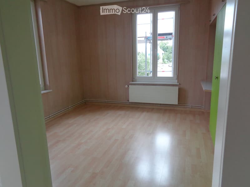 Empty room with wooden flooring, light-colored walls, and a window with white frames. A radiator is visible below the window, and the room has a green accent wall on one side.