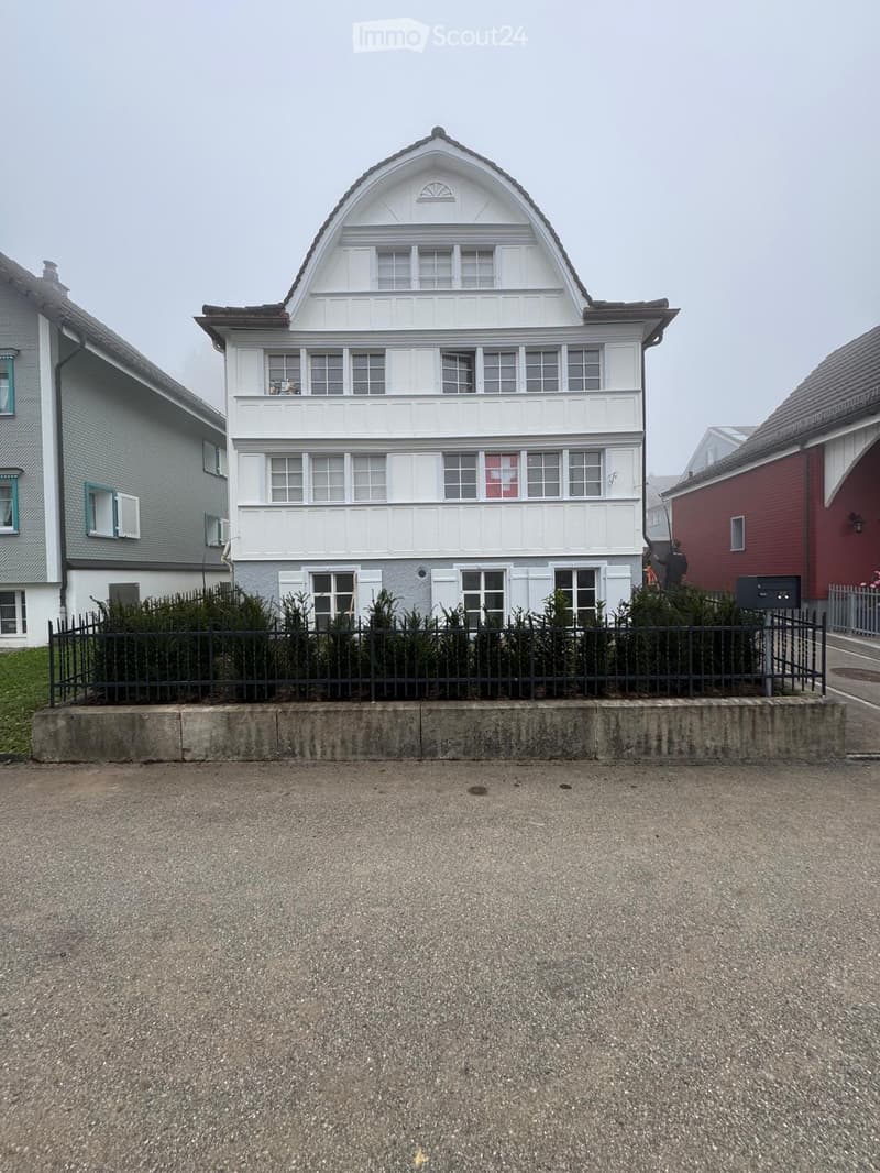 Building with several apartments, white exterior, multiple windows, gray neighboring houses, black fencing, grassy area, road