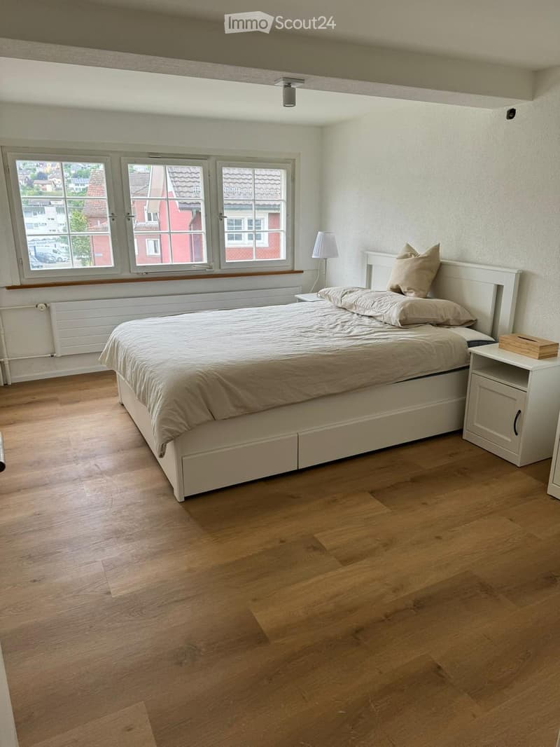 Bedroom with large windows, white bed, bedside tables, lamp, wooden floor, white walls