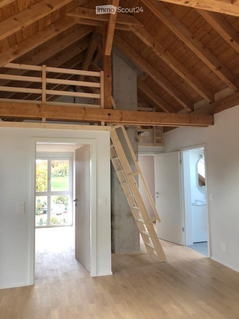 Bright, open loft space with wooden floors, exposed wooden beams, and a wooden staircase leading to an upper level. The room features white walls, large windows, and a simple, clean design.