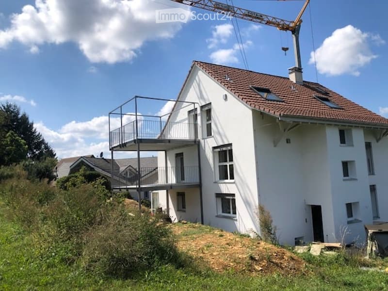 Modern two-story house with white exterior, red-tiled roof, and a raised ground floor. Features a balcony on the upper level and a construction crane in the background.